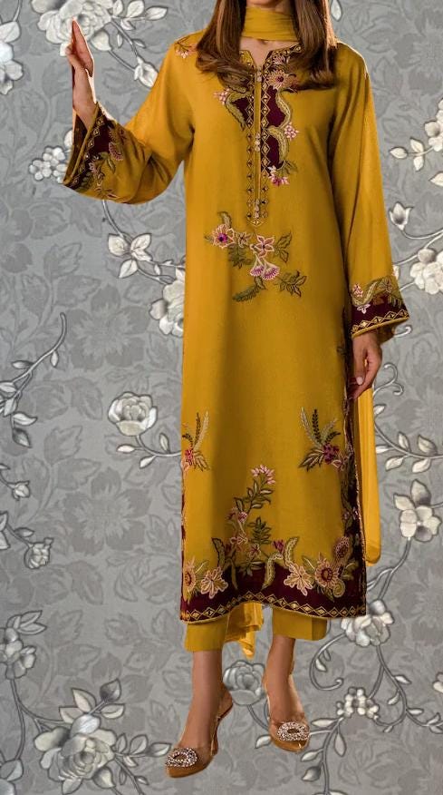 A woman wearing a long, yellow dress with intricate floral embroidery stands against a gray floral background.