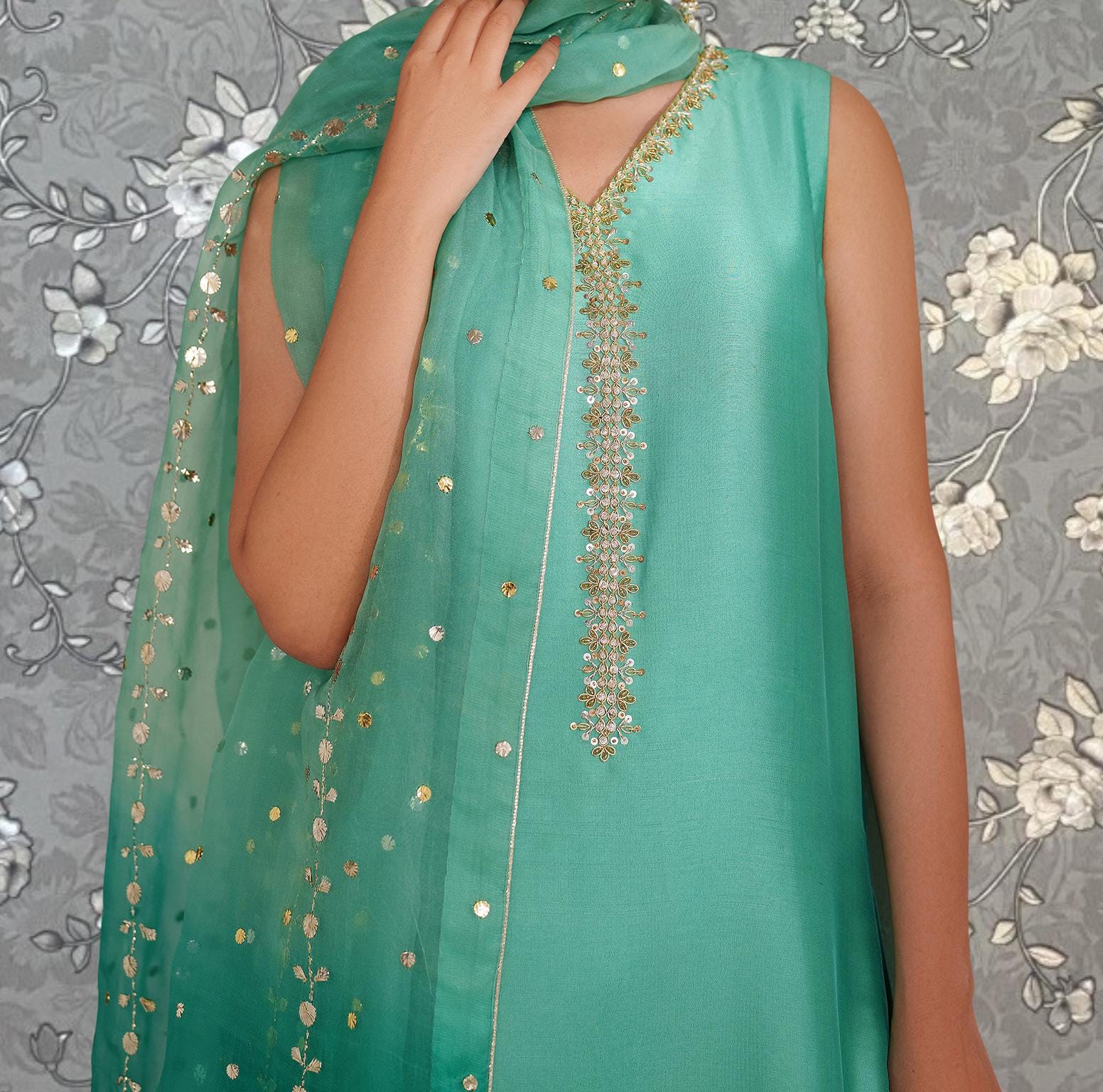 A person wearing a turquoise dress with a green and gold embroidered pattern on the neckline and a matching green and gold scarf draped over the shoulder.