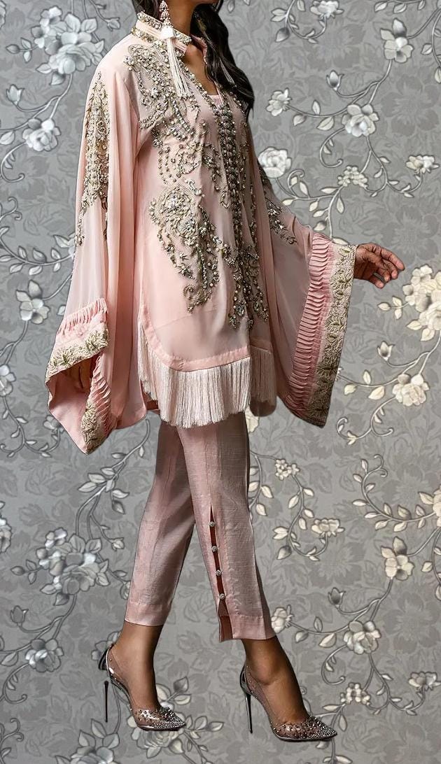 A woman wearing a pink dress with intricate embroidery and frills, standing against a gray floral background.