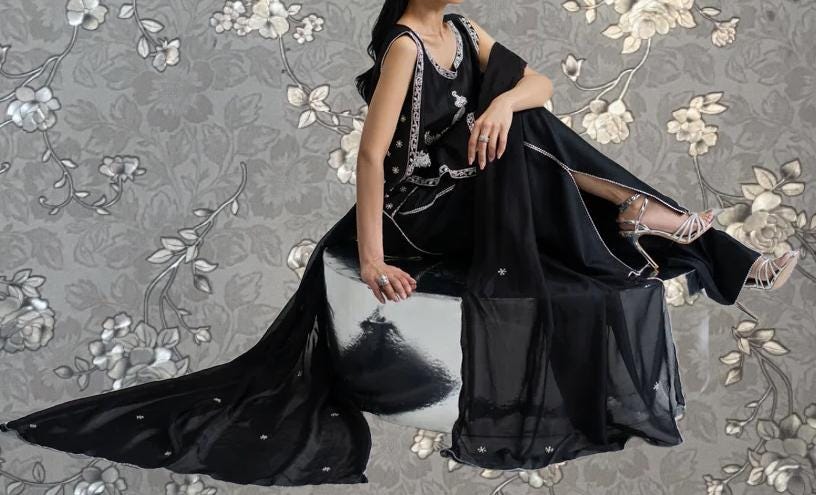 A woman wearing a long black dress with a floral pattern sits on a white and black patterned chair against a gray floral background.