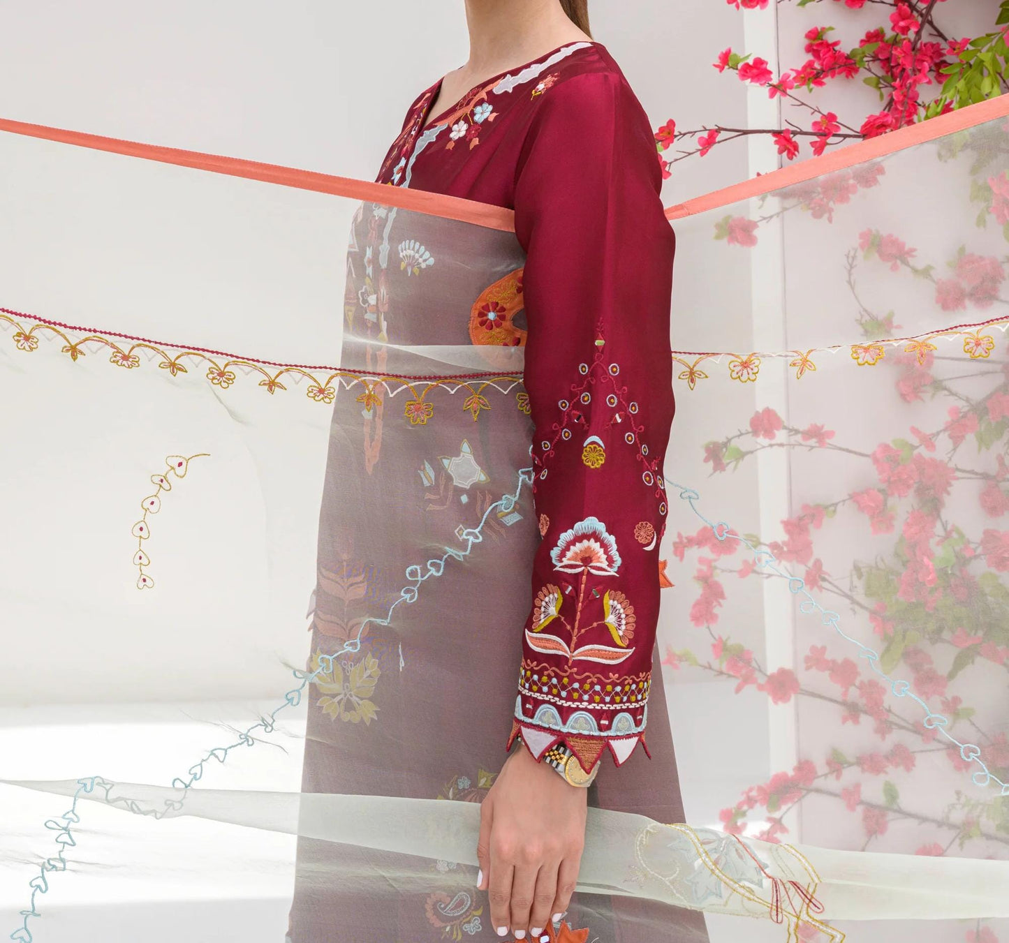 A person wearing a red and white patterned dress stands in front of a white wall with pink flowers.