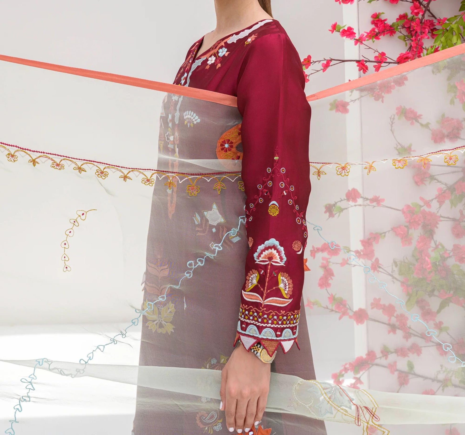 A person wearing a red and white patterned dress stands in front of a white wall with pink flowers.