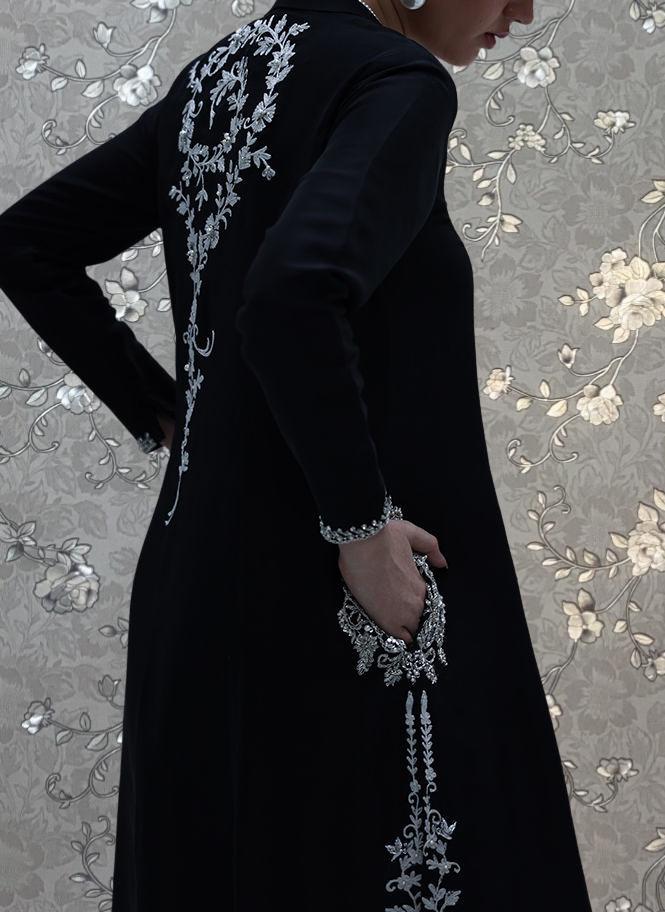 A person wearing a long black dress with intricate white embroidery stands against a floral patterned background.