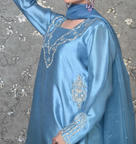 A woman wearing a blue dress with intricate embroidery and jewelry poses against a floral background.