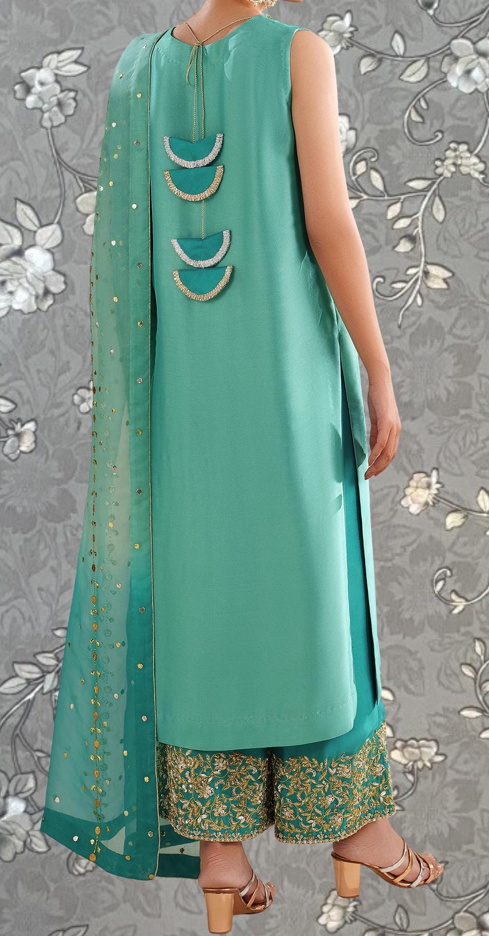 A person wearing a turquoise dress with gold embellishments and gold heels, standing against a gray floral background.