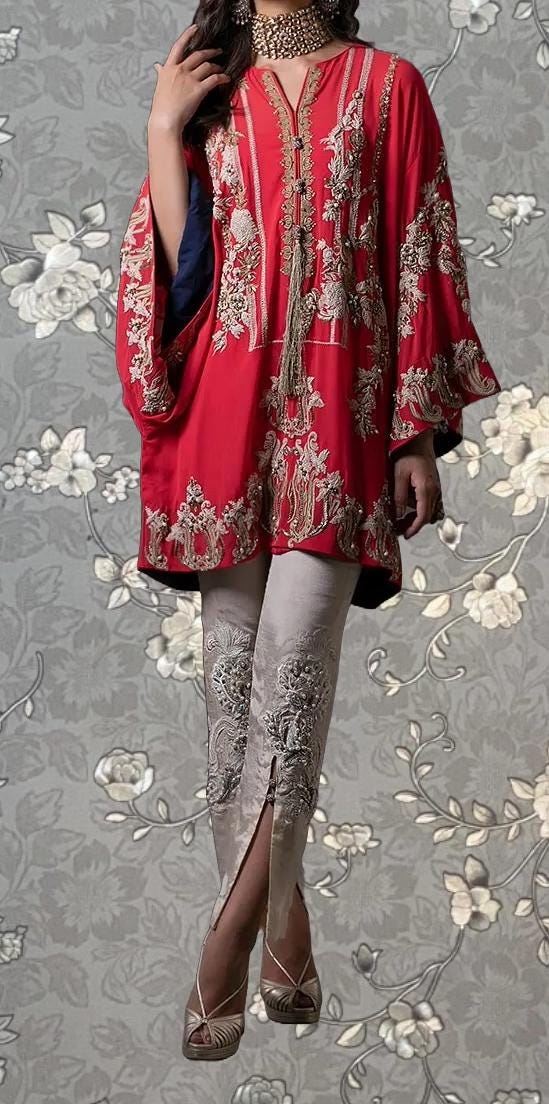 A woman wearing a red and gold embroidered dress with a high slit on the left leg, standing against a gray floral background.