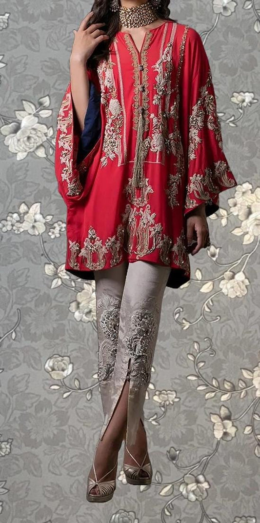 A woman wearing a red and gold embroidered dress with a high slit on the left leg, standing against a gray floral background.