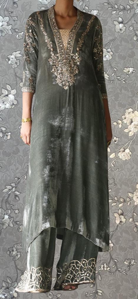 A person wearing a gray and gold dress with intricate embroidery and patterns, standing against a gray floral background.