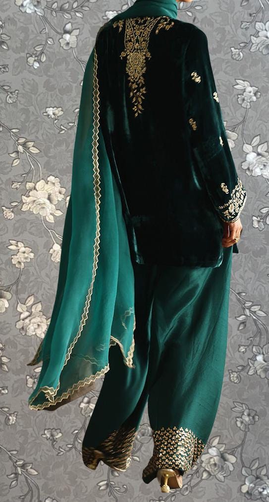 a person wearing a green and black outfit with gold embroidery, standing against a gray floral background.