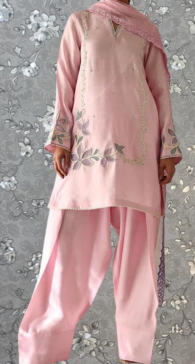 A person wearing a pink dress with floral embroidery stands against a gray floral background.