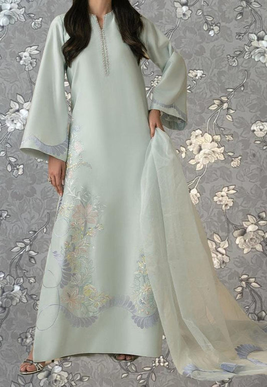 A woman wearing a long, light blue dress with intricate floral embroidery stands against a gray floral background.