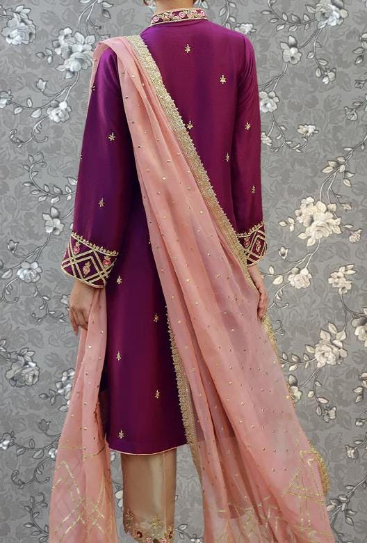 a person wearing a purple and pink dress with a pink shawl draped over the shoulders. The dress has intricate embroidery and gold accents, and the person is standing against a gray floral background.