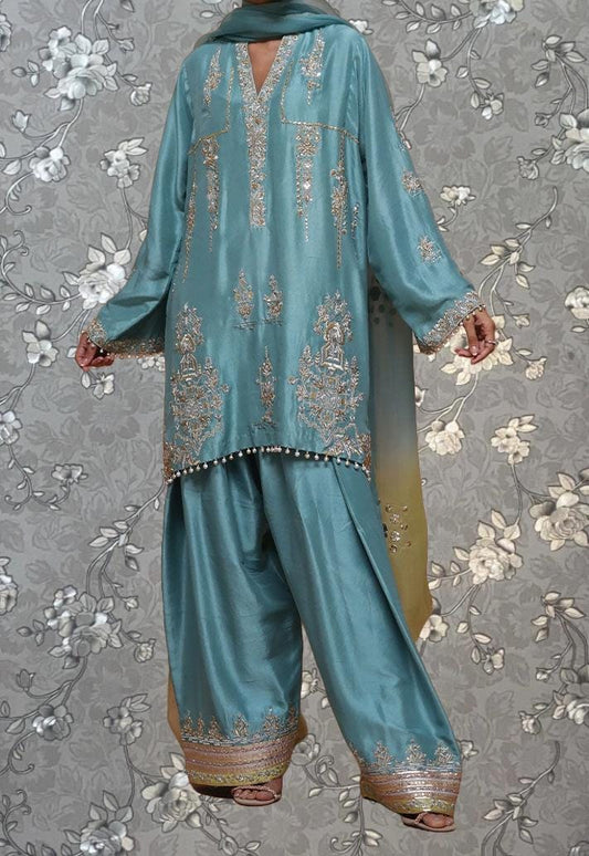 A person wearing a turquoise and gold embroidered dress stands against a gray floral background.