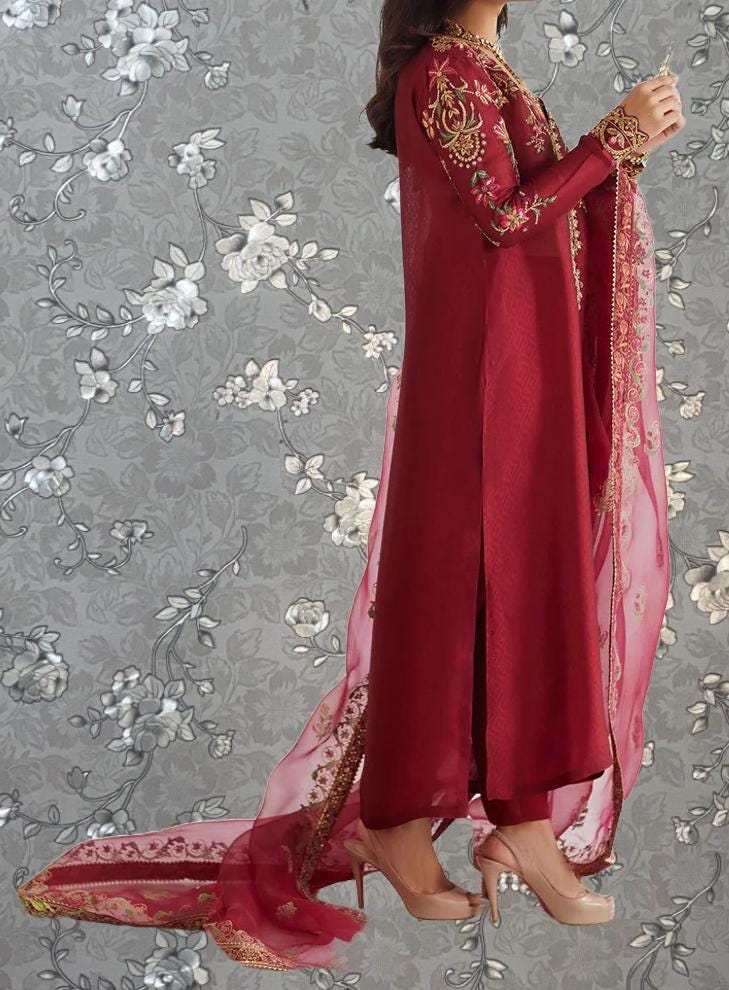 A woman wearing a long, red dress with intricate embroidery stands against a gray floral background.
