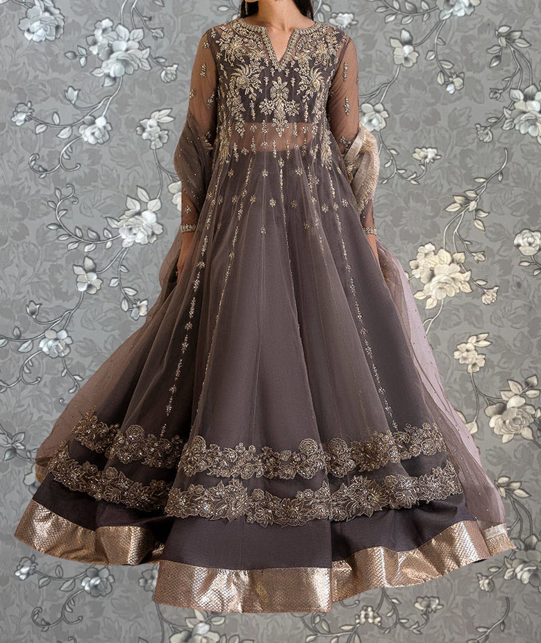 A woman wearing an ornate, long dress with intricate embroidery and gold accents, standing against a gray floral background.