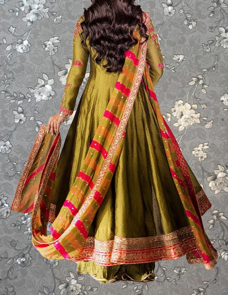 A woman wearing a long, green dress with a red and yellow patterned skirt, holding a sash, and standing against a gray floral background.