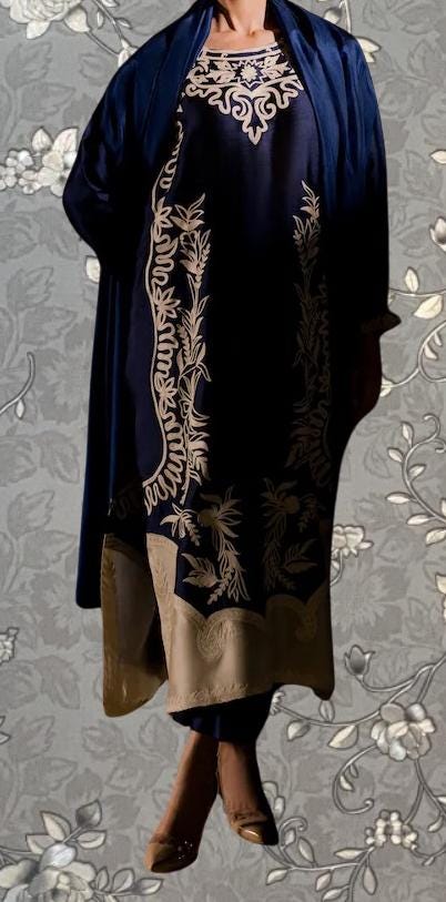 A person wearing a navy blue dress with gold embroidered patterns, standing against a gray floral background.