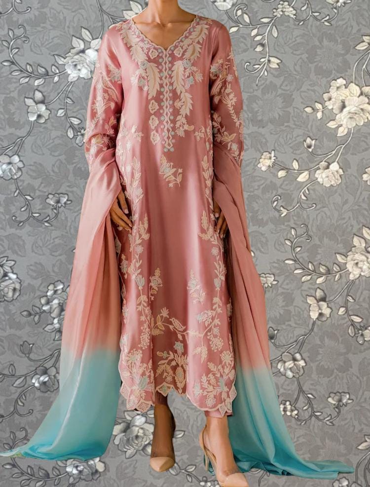 A woman wearing a pink dress with intricate floral embroidery and a matching pink shawl stands against a gray floral background.