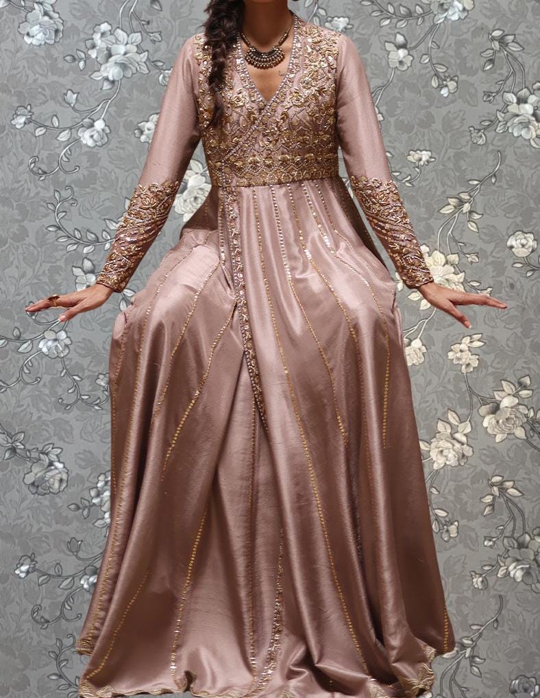 A woman wearing a long, pink dress with intricate gold embroidery and a high neckline stands against a gray floral background.