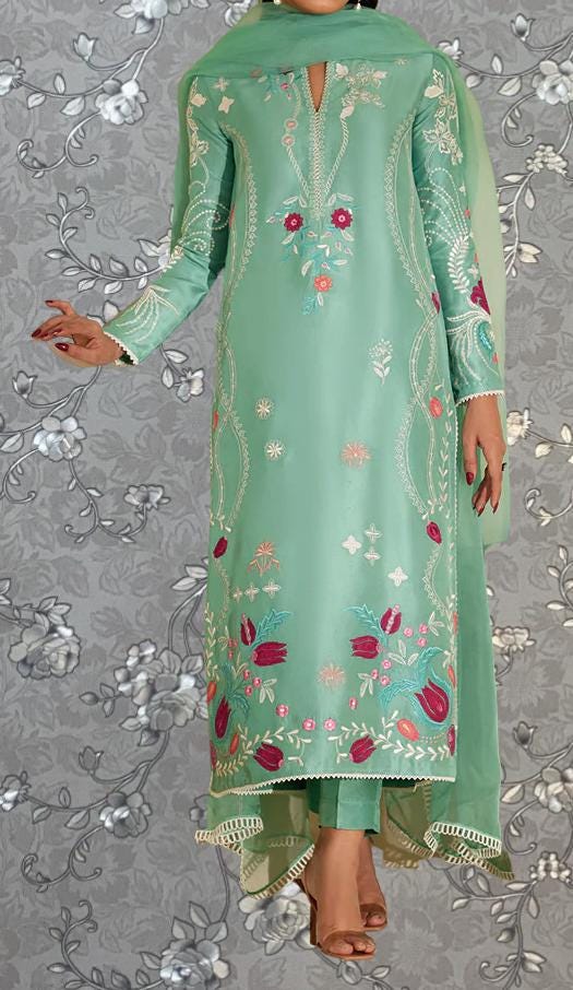 A woman wearing a long, green dress with intricate floral embroidery stands against a gray floral background.