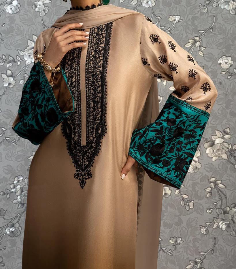 A person wearing a beige dress with intricate embroidered patterns in black and teal colors, standing against a floral background.