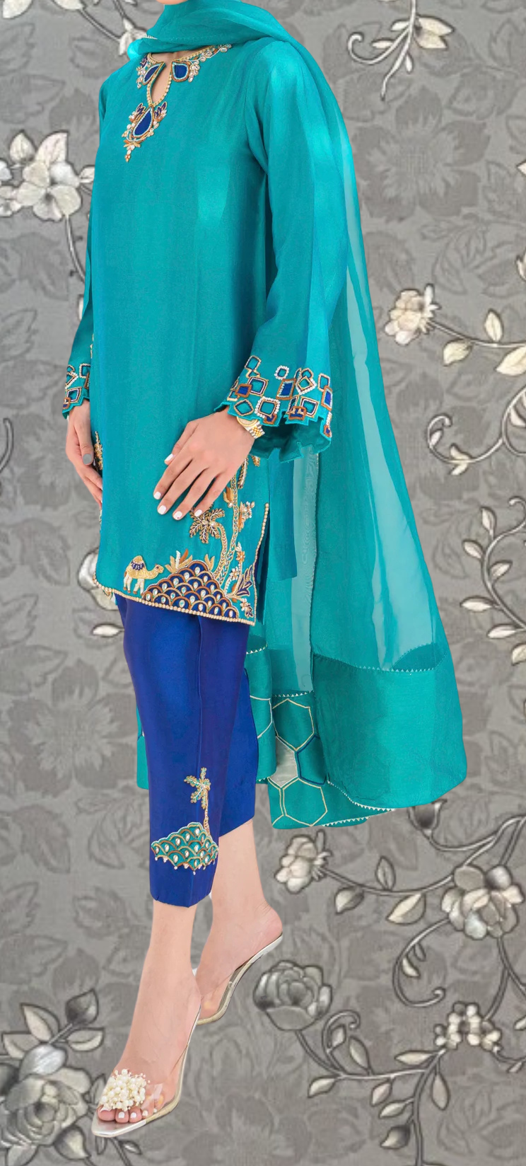 A person wearing a turquoise dress with intricate embroidery and blue pants stands against a gray floral background.