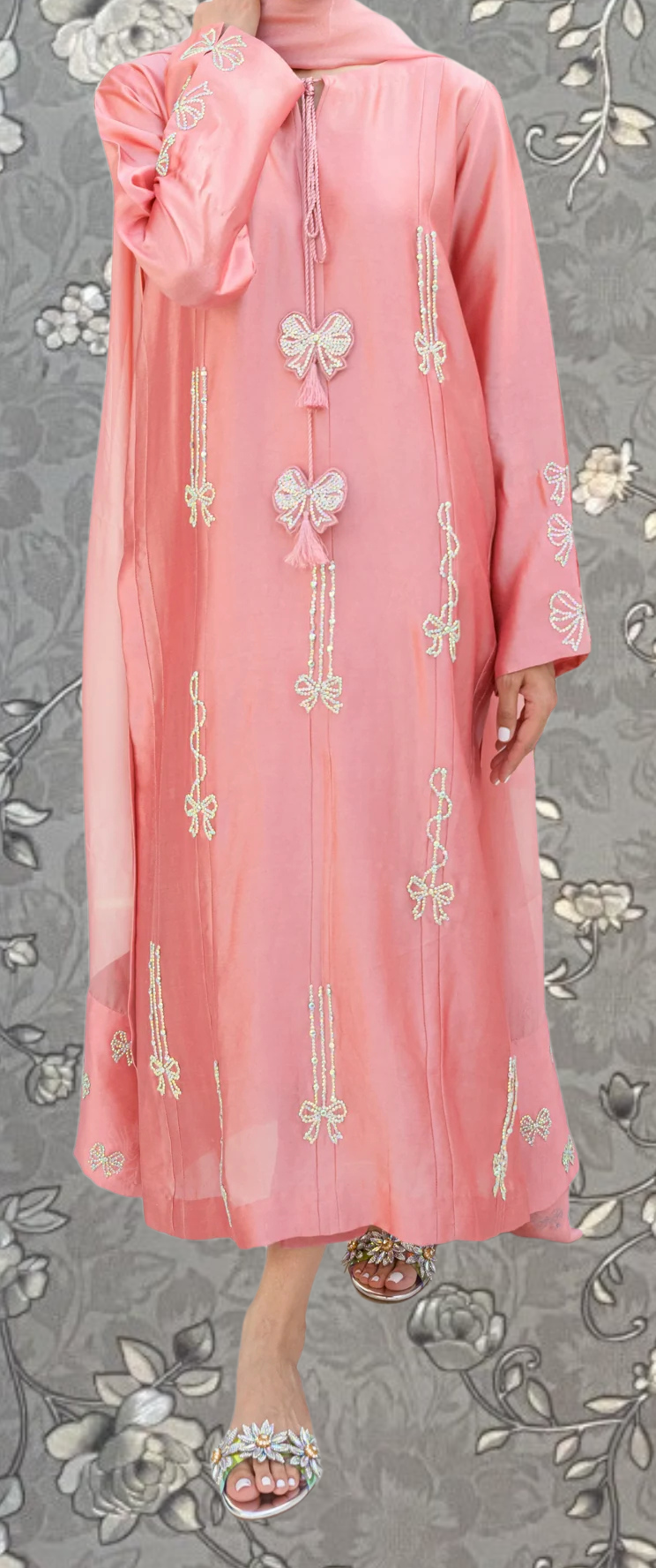 A woman wearing a pink dress with white floral embroidery and tassels, standing against a gray floral background.