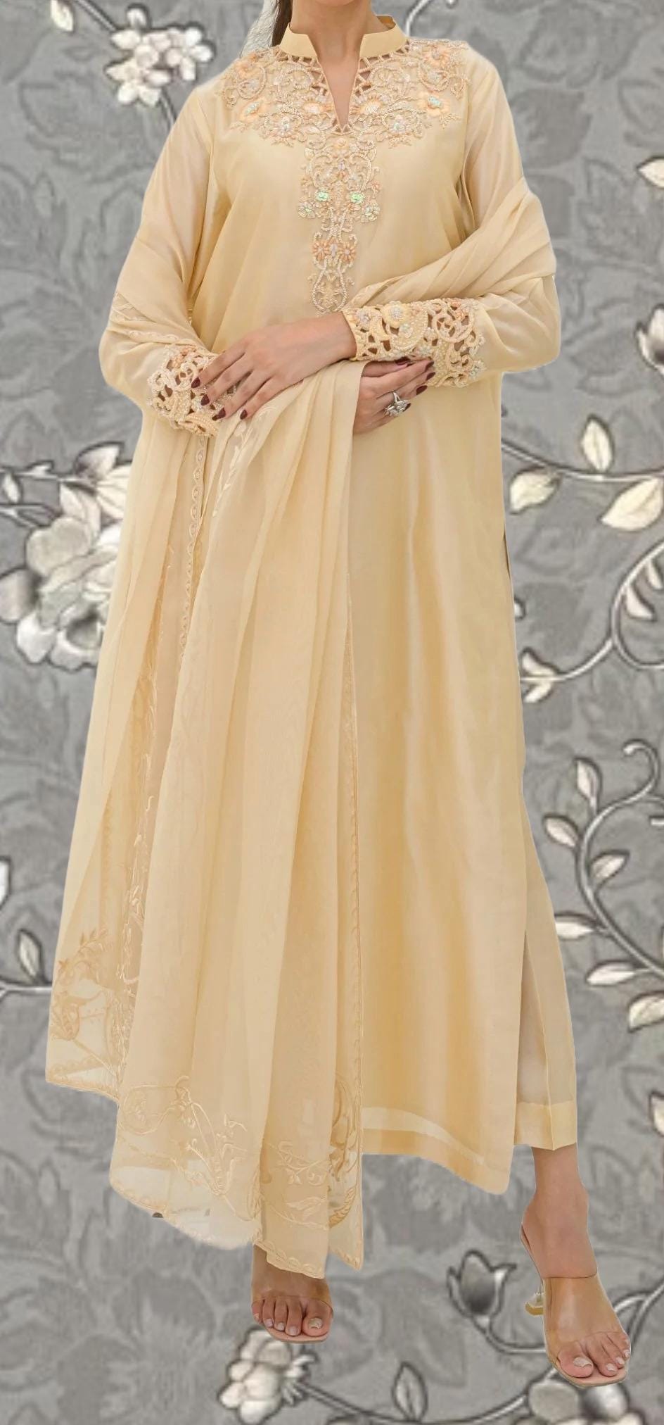 A woman wearing a long, cream-colored dress with intricate embroidery and lace details, standing against a gray floral background.