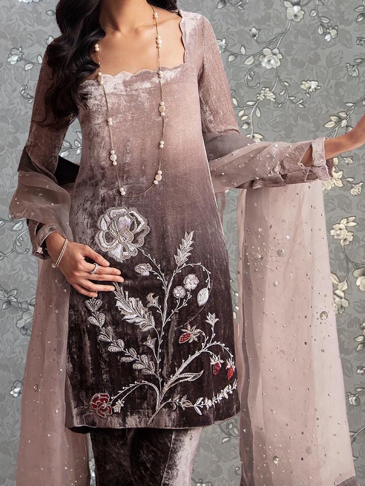 A woman wearing a long, beige dress with intricate floral embroidery stands against a gray floral background.