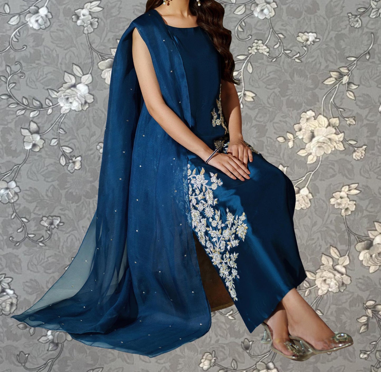 A woman wearing a blue dress with white floral embroidery is sitting against a gray floral background.