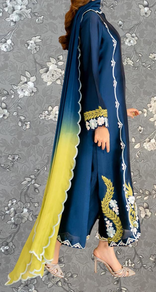 A woman wearing a blue and yellow dress with intricate embroidery and a matching yellow shawl, standing against a gray floral background.