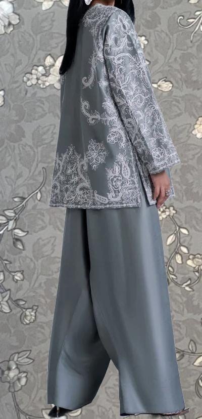 A person wearing a gray and white patterned outfit with intricate embroidery stands against a floral background.