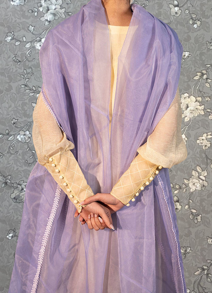 A person wearing a purple and yellow dress with a long purple shawl draped over their shoulders, standing against a gray floral background.