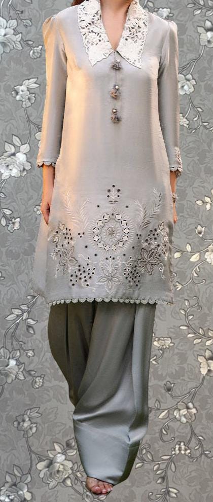 A woman wearing a gray dress with intricate embroidery and a white collar, standing against a gray floral background.