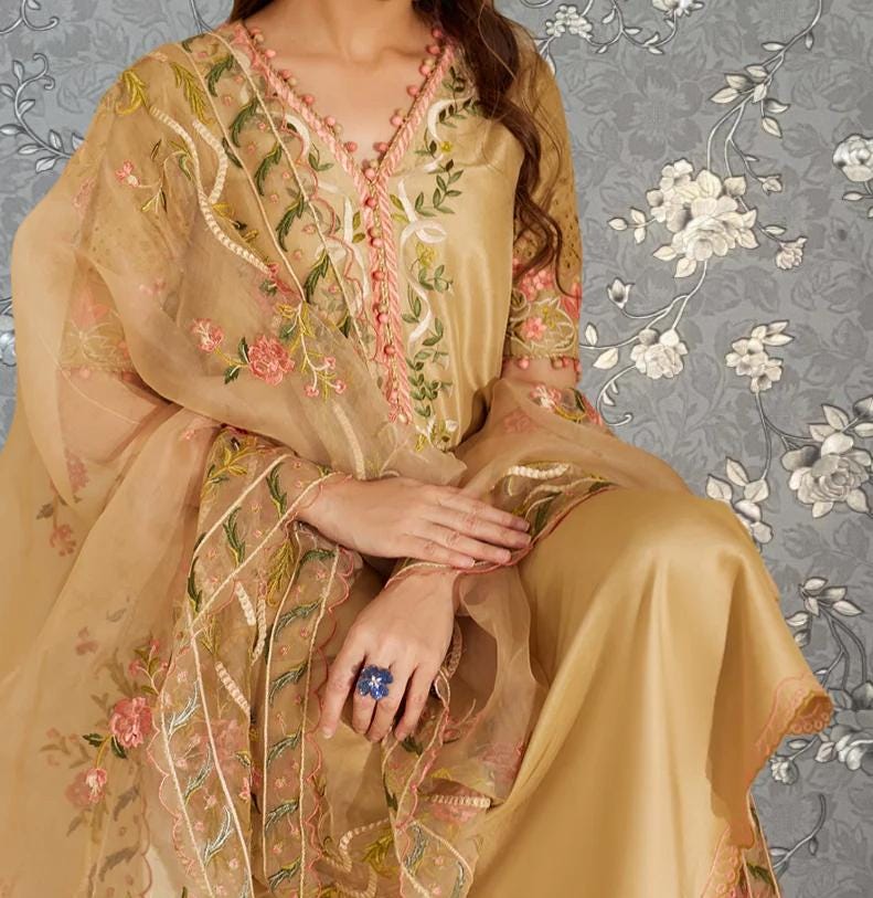 A woman wearing a gold and beige dress with intricate floral embroidery and a matching shawl. She is holding a ring on her finger.