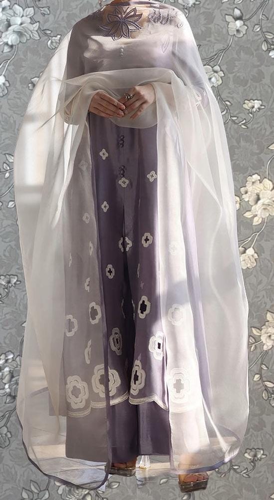 A person wearing a long, flowing purple dress with white floral patterns, standing against a gray floral background.