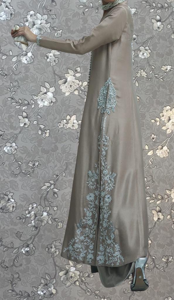 A person wearing a long, beige dress with intricate floral embroidery stands against a gray floral background.