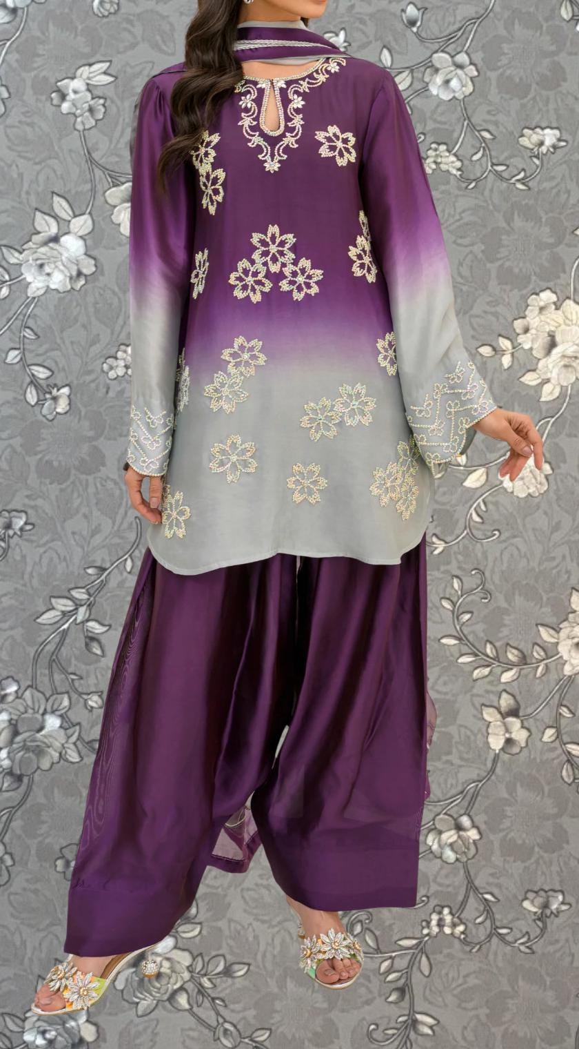 A woman wearing a purple and gray dress with white floral embroidery, standing against a gray floral background.