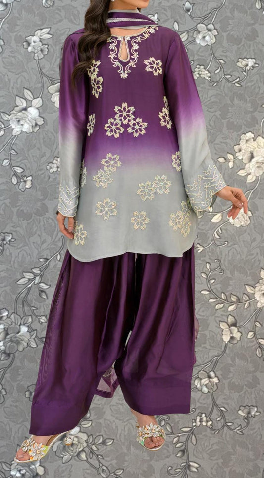 A woman wearing a purple and gray dress with white floral embroidery, standing against a gray floral background.
