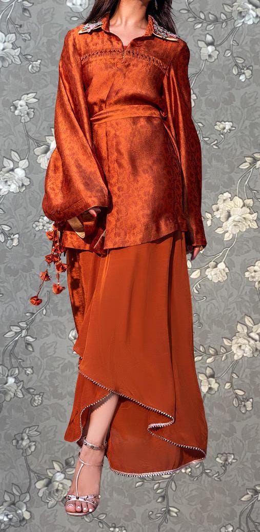 A woman wearing a long orange dress with a high collar and long sleeves, standing against a gray floral background.