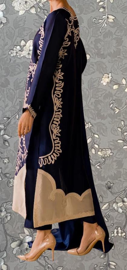 A person wearing a long, dark blue dress with intricate white embroidery stands against a gray floral background.