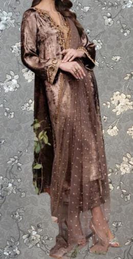 A woman wearing a long, brown velvet dress with a sheer, polka-dotted overlay. She is posing with her hands on her hips, against a gray floral background.