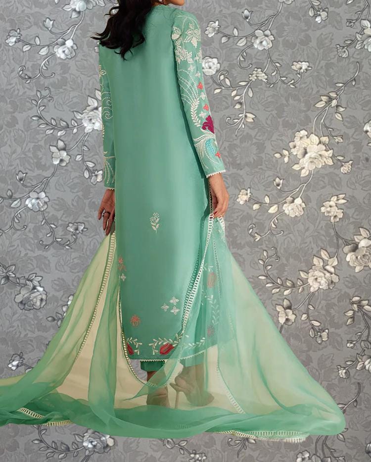 A woman wearing a long, mint green dress with floral embroidery stands against a gray floral background.