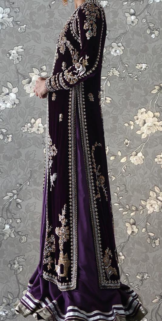 A person wearing an ornate purple and gold dress with intricate embroidery and patterns, standing against a gray floral background.