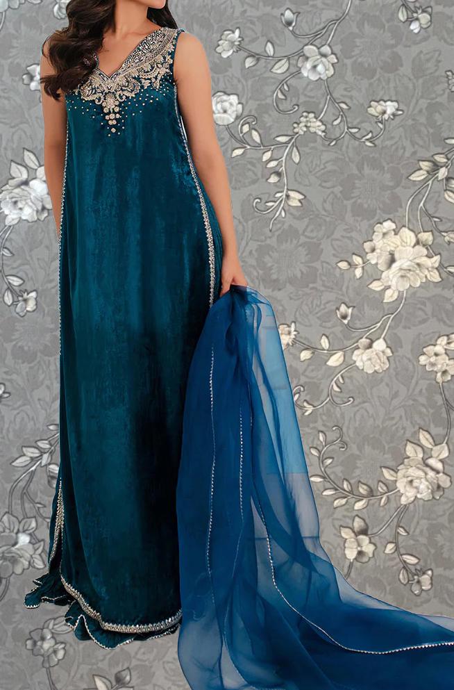A woman wearing a long, teal velvet dress with gold embroidery stands against a gray floral background.