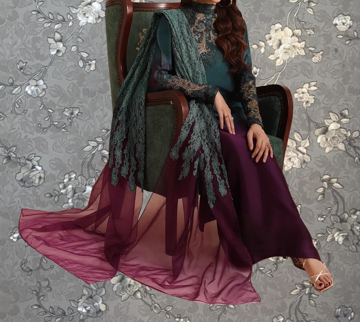 A woman wearing a long, flowing green and purple dress with intricate embroidery, sitting in a chair with a floral patterned background.
