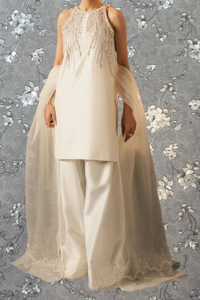 A woman wearing a white dress with beaded embellishments and a sheer white shawl, standing against a gray floral background.