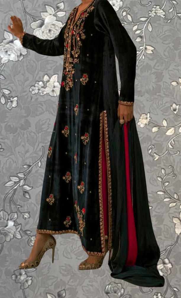 A person wearing a long black dress with red and gold embroidered patterns, standing against a gray floral background.