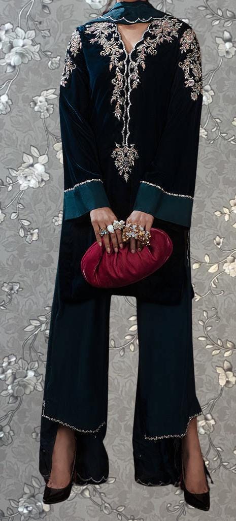 A person wearing a long black dress with intricate embroidery and holding a red clutch purse stands against a floral patterned background.