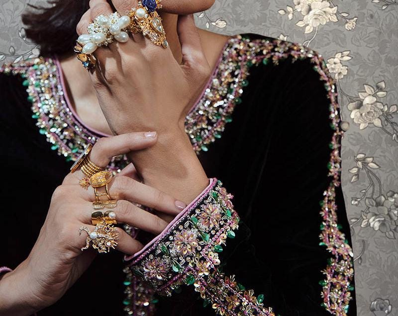 A close-up of a person's hand adorned with an array of jewelry, including multiple rings, bracelets, and necklaces, against a floral patterned background.